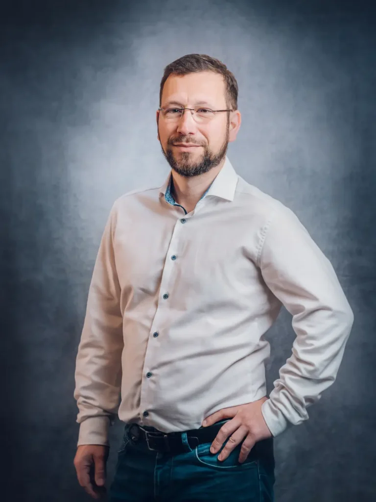 Dipl.-Ing. (FH) Thomas Wagner steht selbstbewusst mit einer Hand in der Hüfte da. Er trägt ein helles Hemd und Jeans vor einem grauen, strukturierten Hintergrund.