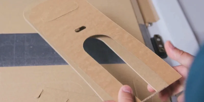 Nahaufnahme einer Person, die ein ausgeschnittenes Stück Pappe hält, das einen rechteckigen Ausschnitt mit einer ovalen Aussparung zeigt. Im Hintergrund ist weitere Pappe zu sehen.